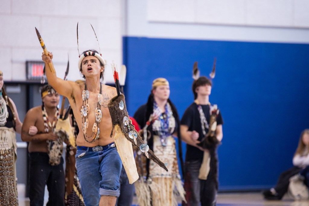 A group of dancers performing in traditional attire, featuring a shirtless man leading the performance while holding a spear and wearing a feathered headdress, surrounded by others in ceremonial clothing.