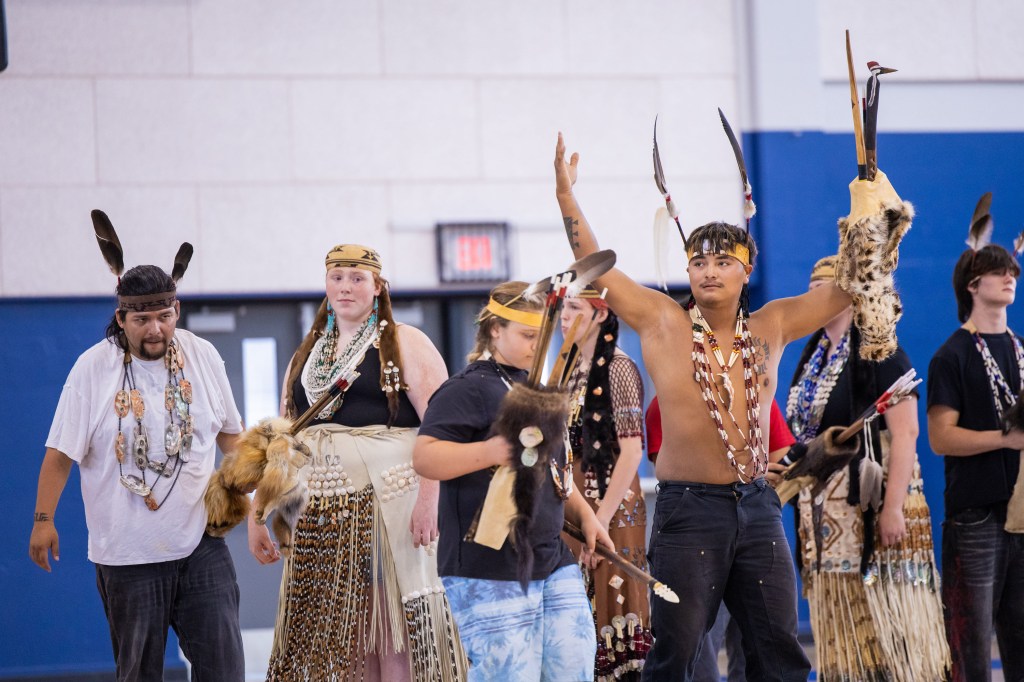 A group of Indigenous dancers dressed in traditional attire performing in a gymnasium, showcasing various accessories such as feathers, beads, and animal skins.