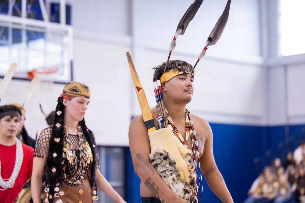 A young Indigenous man dressed in traditional attire, carrying a bundle and adorned with feathers and beads, participates in a cultural event alongside others in similar clothing.