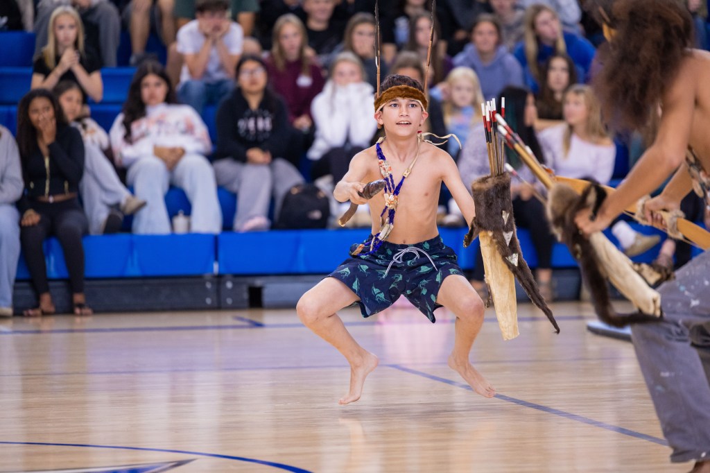 A young boy in traditional attire performs a dance in a gymnasium, with spectators watching in the background.