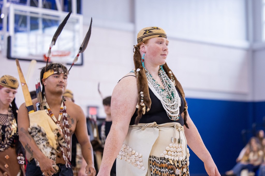 A group of individuals participating in a traditional cultural dance, dressed in ornate costumes with feathers and beadwork. The scene captures a male dancer in the foreground holding a traditional item, while a female dancer, adorned with jewelry and intricate attire, leads the group.