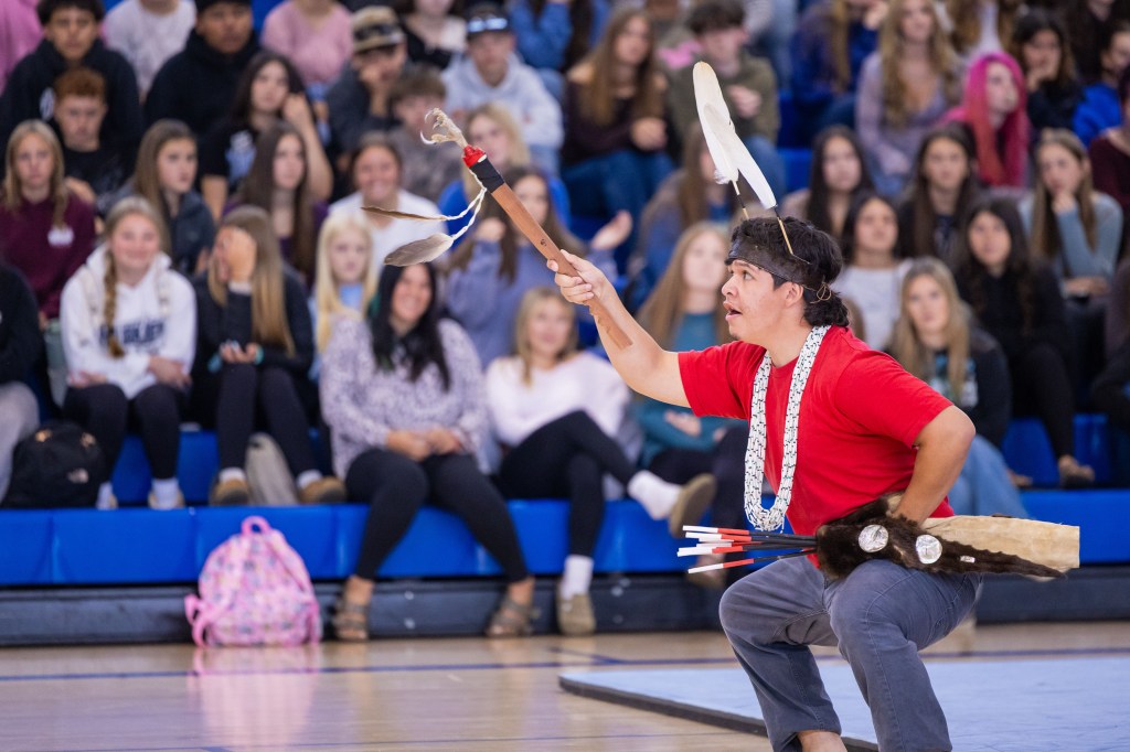 A performer in a red shirt and traditional attire showcases a dance while holding a decorated pole with feathers in a gymnasium, with an audience in the background.