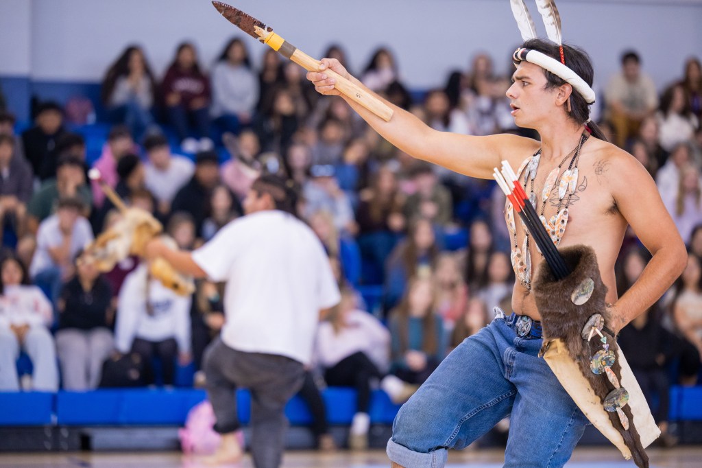A performer dressed in traditional attire, holding a spear and wearing tribal accessories, showcases a cultural dance in front of an attentive audience.