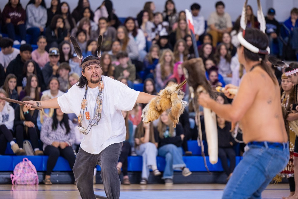 A performer in traditional attire engages in a cultural dance, displaying a ceremonial object while surrounded by an audience in a gymnasium.