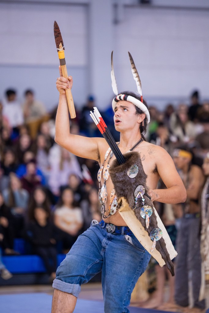 A young man performing in traditional attire, holding a spear and wearing a feathered headdress, in front of a seated audience.