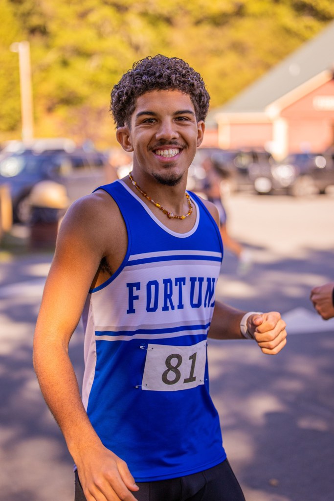 Smiling young male athlete wearing a blue tank top with 'FORTUNE' printed on it and a race number, preparing for a run outdoors.