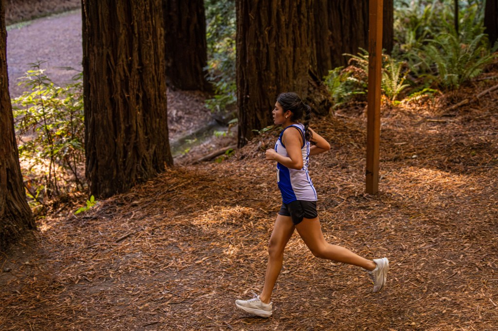 A young woman jogging on a dirt trail surrounded by tall trees and greenery.