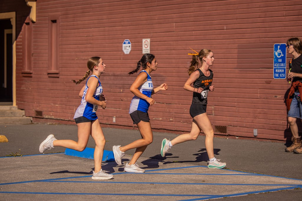 Three female athletes running in a race on a track, with a red building in the background.