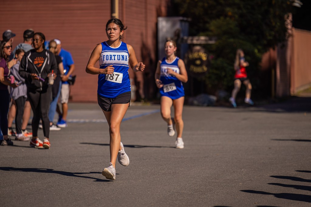 Two female runners in blue athletic uniforms competing in a race, with spectators watching in the background.