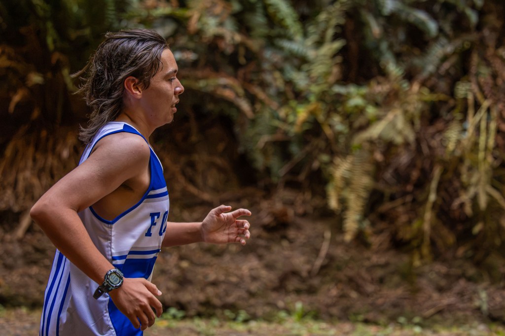 A young athlete running on a forest trail, wearing a blue and white sports jersey and a watch.