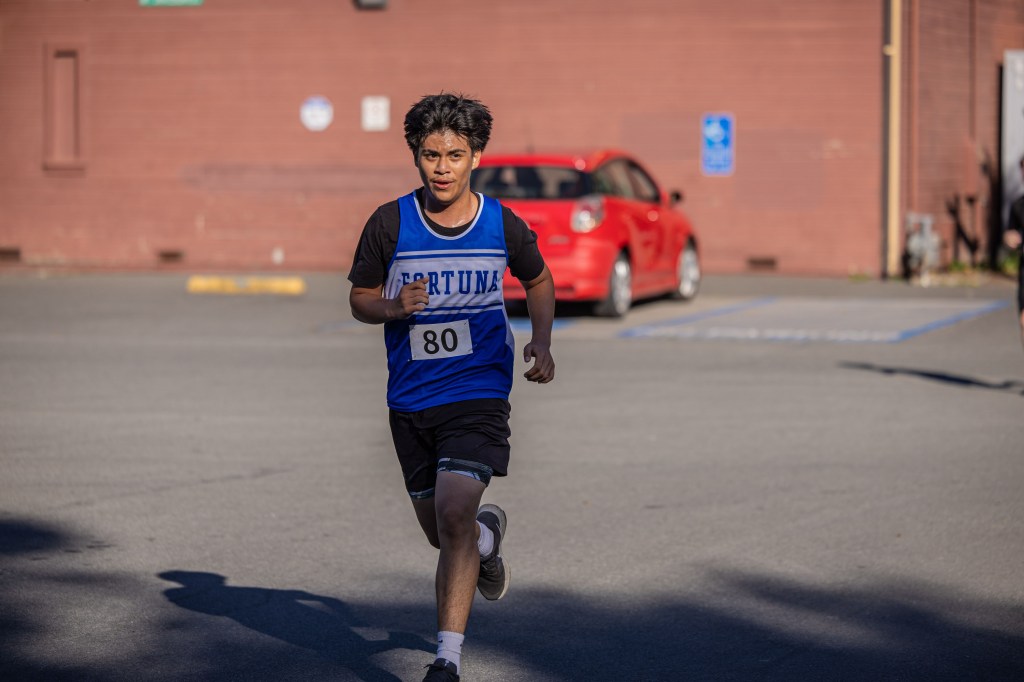 A young runner in a blue sports jersey with the number 80 is sprinting on a parking lot with a red car in the background.