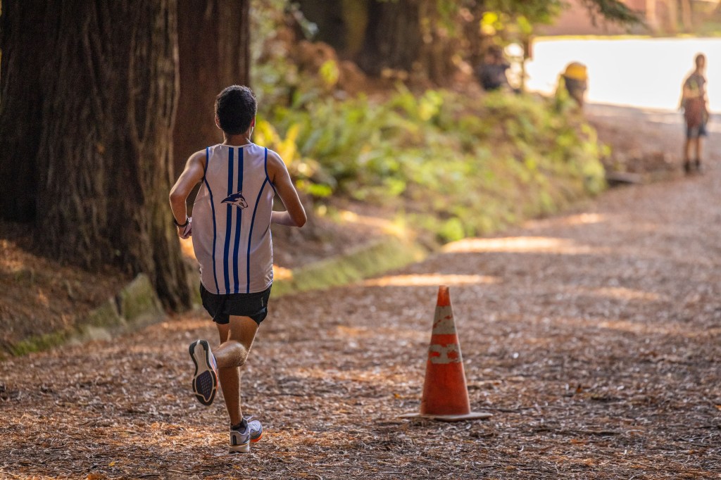 A young male runner in a tank top and shorts jogging down a forested path, with a traffic cone visible on the side of the trail.