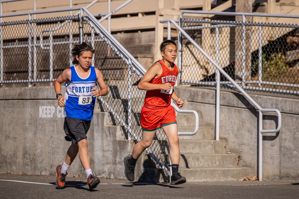 Two young male runners competing in a race, one wearing a blue jersey with 'FORTUNE' and the number 83, the other in a red jersey labeled 'EUREKA'.