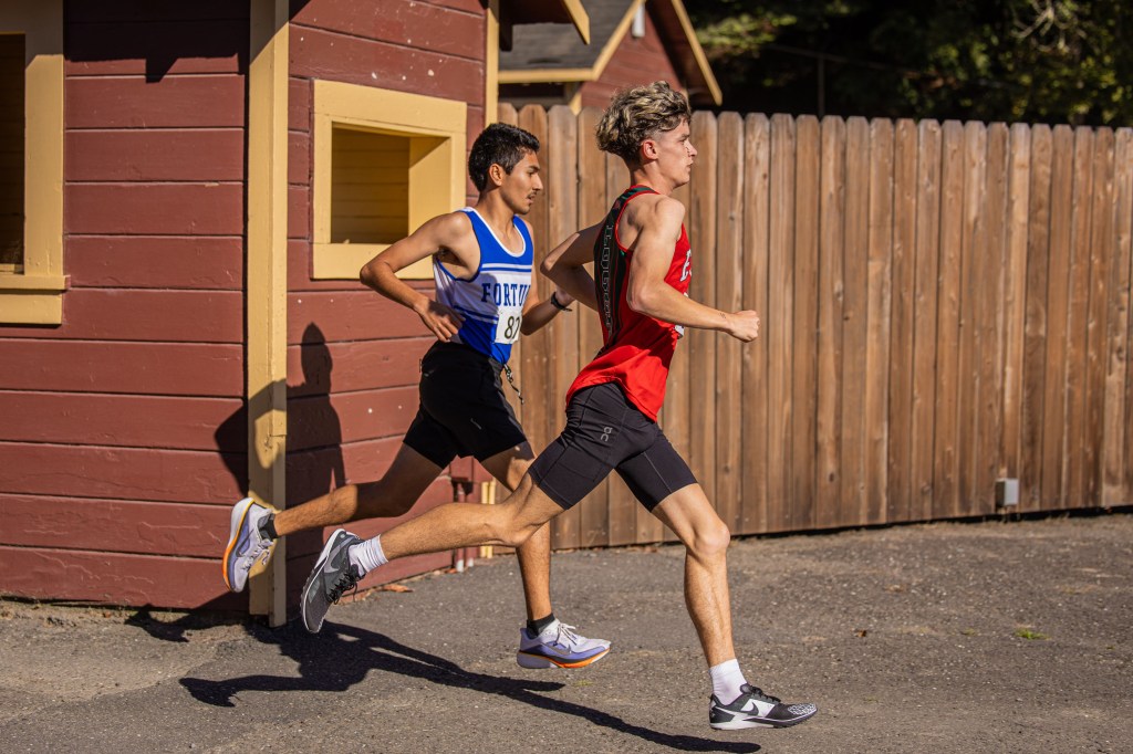 Two male runners in athletic wear sprinting on a track, one in a blue jersey and the other in a red jersey, with a wooden fence in the background.