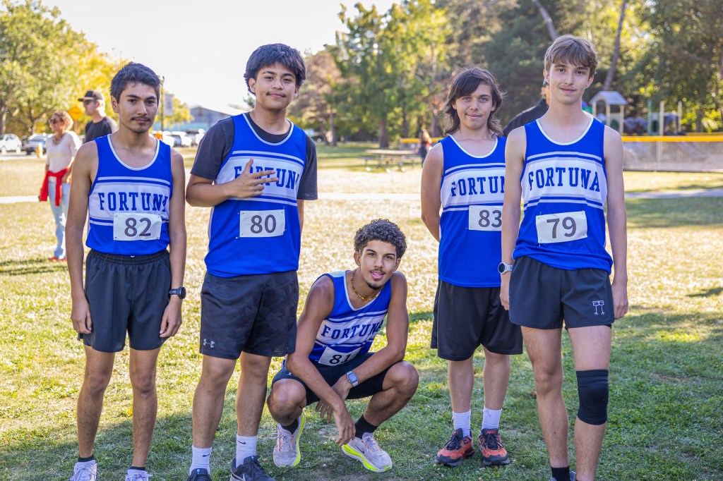 Five male cross country runners posing together in blue and white jerseys with the text 'FORTUNA' on them, standing on a grassy field.