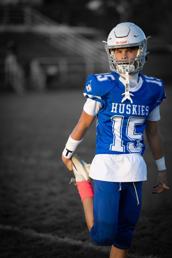 A young football player in a blue jersey with 'Huskies' printed on it, stretching his leg while wearing a helmet and black cleats on a football field.