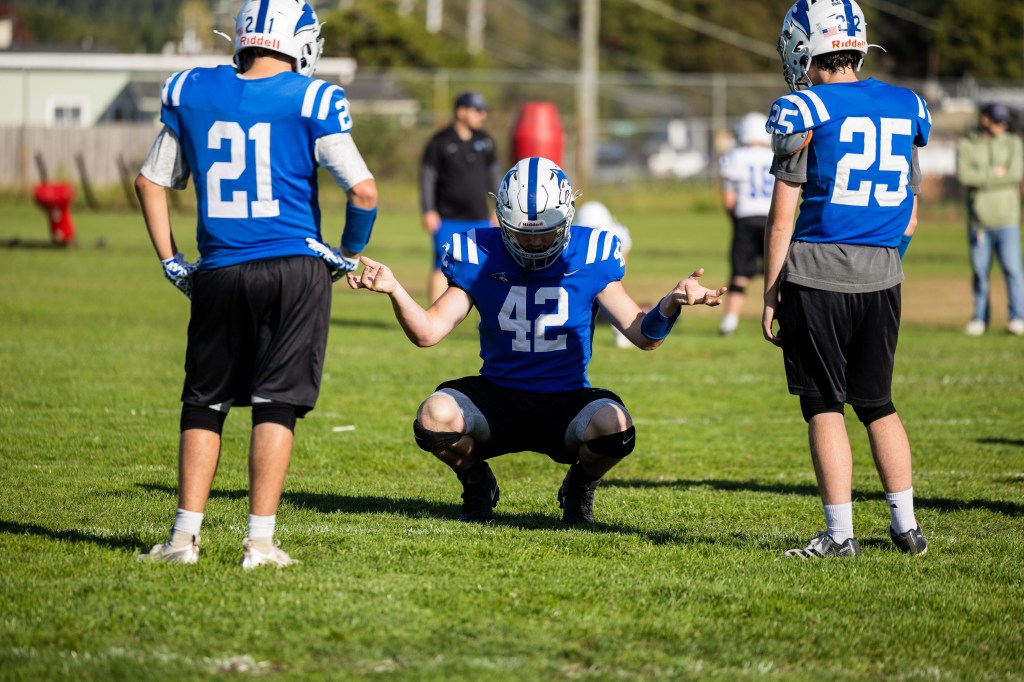 Three football players on a field; one player in a blue jersey squats with hands raised, while two teammates stand nearby.