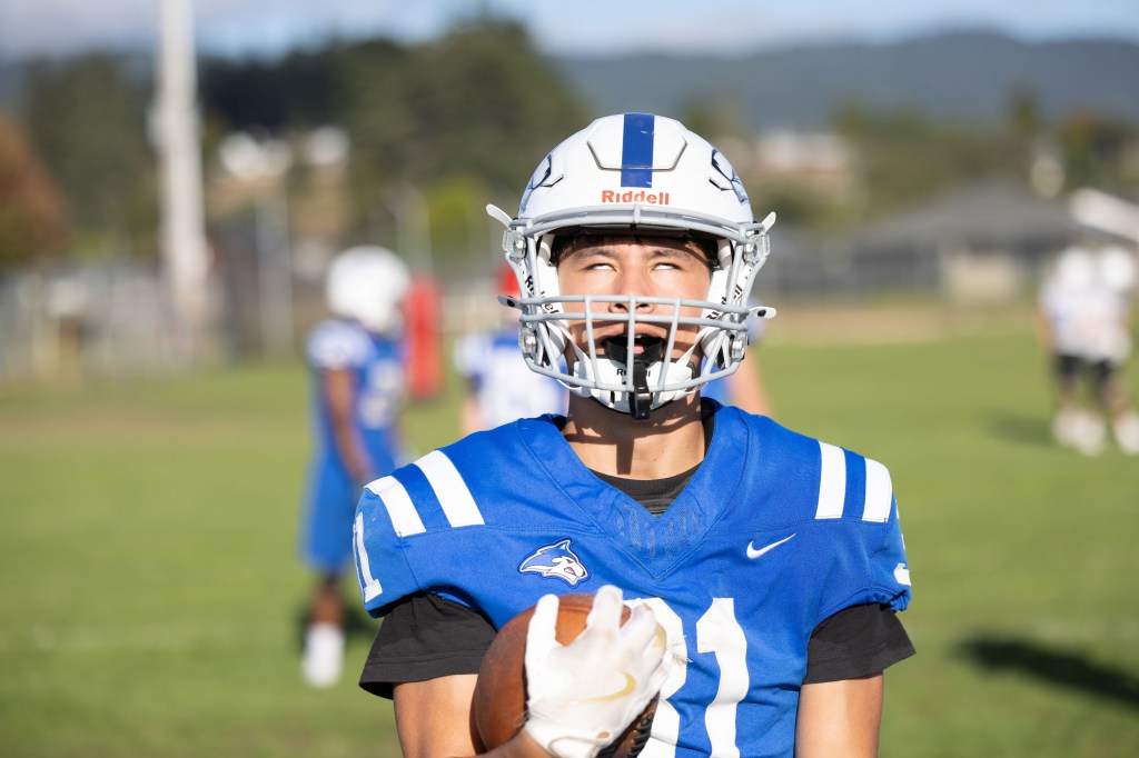 A young football player wearing a blue jersey and a helmet, holding a football and smiling, surrounded by teammates on a grassy field.