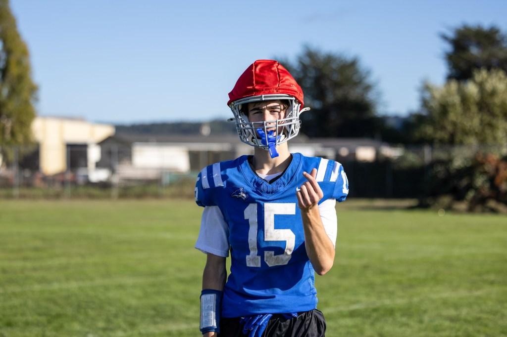 A young male football player wearing a blue jersey with the number 15 and a red helmet, standing on a grassy field and giving a playful hand gesture.