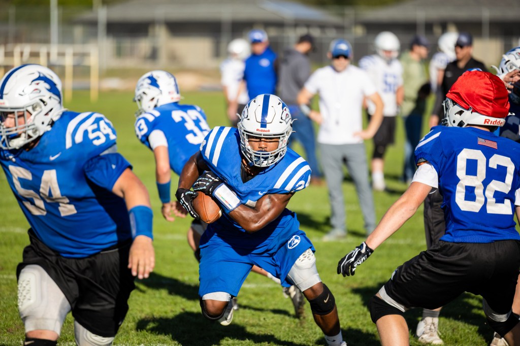 A football player in blue jersey and helmet is running with the ball on the field, while teammates and opponents are moving in the background.