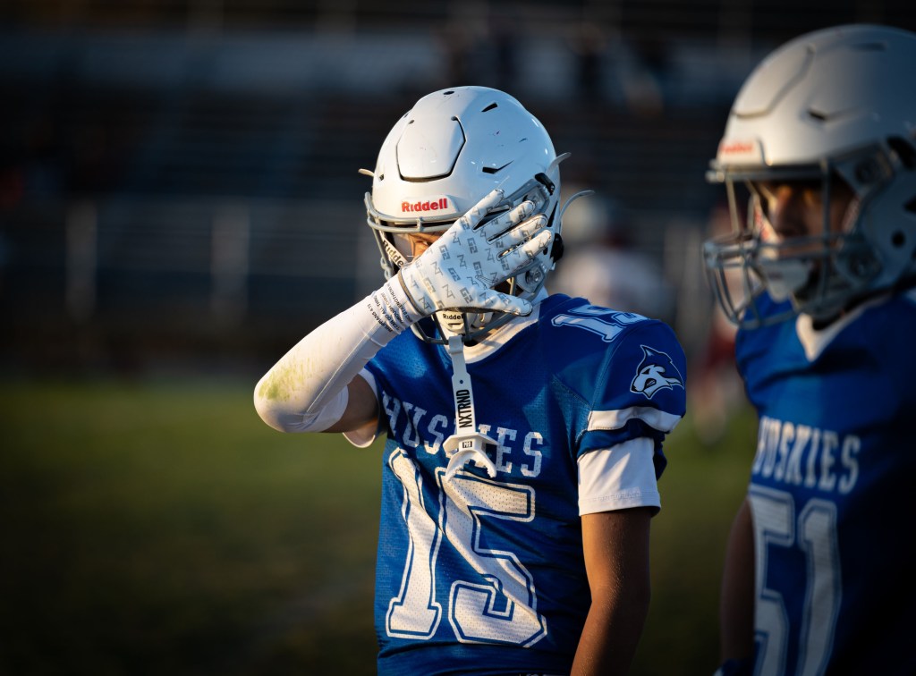A youth football player in a blue jersey with the number 15 stands on the field, partially covering his face with his hand. Another player in a blue jersey is positioned nearby.
