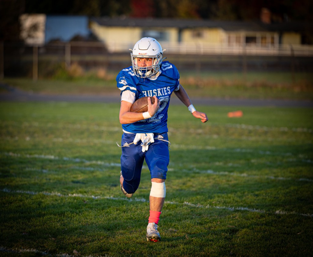 A football player in a blue uniform with 'Huskies' written on it is running on a grass field while holding a football. The player wears a white helmet and has one pink sock visible.