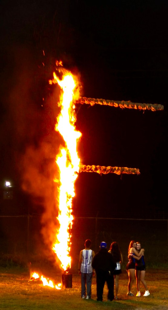 A large flaming structure in the shape of a cross burns brightly at night, with several people standing nearby, some embracing and others watching.