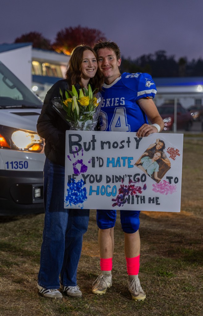 A young couple poses together at a football event, smiling and holding a sign that invites the girl to homecoming, while the boy wears a blue football jersey and pink socks.