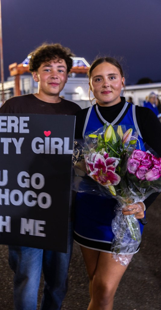 A young couple poses for a photo during an event, with a boy holding a sign that says 'You're a pretty girl, will you go to homecoming with me?' and a girl in a cheerleading outfit holding a bouquet of flowers.