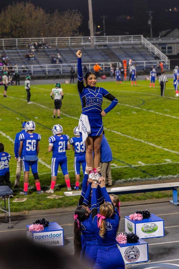 A cheerleader performing a stunt by being held aloft by teammates on a football field, wearing a blue and white uniform, with players in the background.