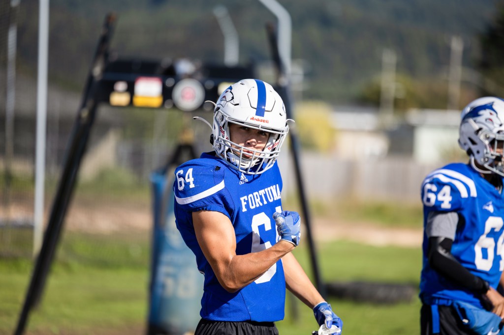 A young football player in a blue jersey with the number 64 flexes his arm, showing off his muscles, during a practice session. Another player in a similar jersey stands nearby, with a background of sports equipment and a field.