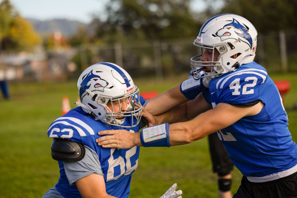 Two football players in blue jerseys practicing a drill on a grassy field, one player blocking while the other pushes forward.