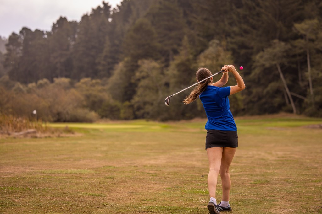 A woman in a blue shirt and black shorts swings a golf club on a grassy course, with trees in the background.