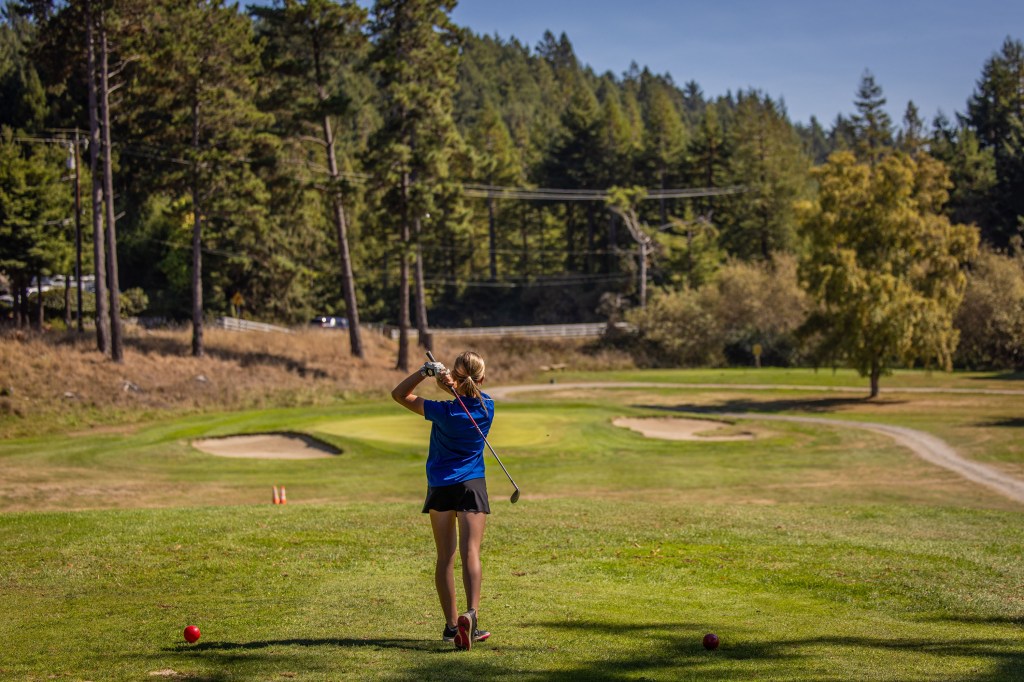 A golfer in a blue shirt and black shorts is preparing to swing a club on a golf course, with trees and a sand trap in the background.