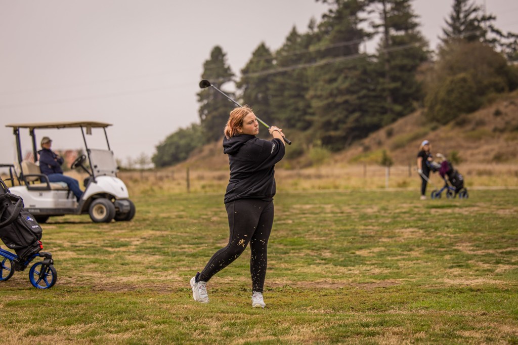 A woman in a black hoodie swings a golf club on a grassy course, with a golf cart and other players in the background.