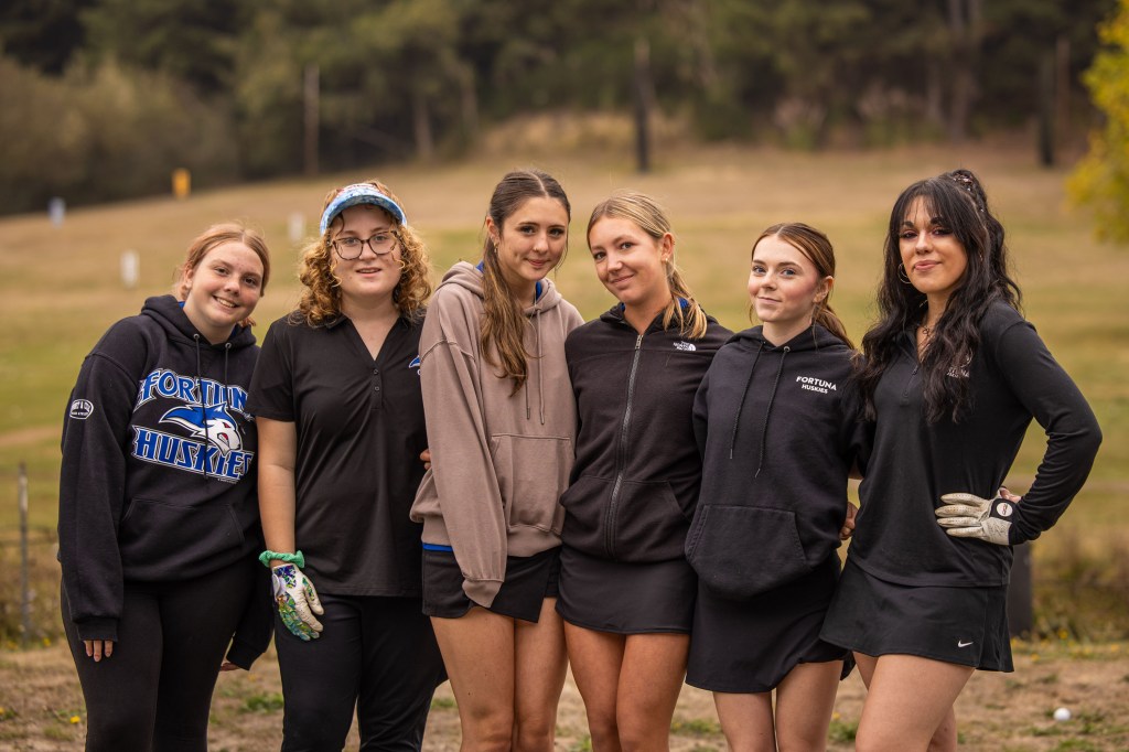 Group of six young women standing together outdoors, all wearing athletic clothing, smiling at the camera.