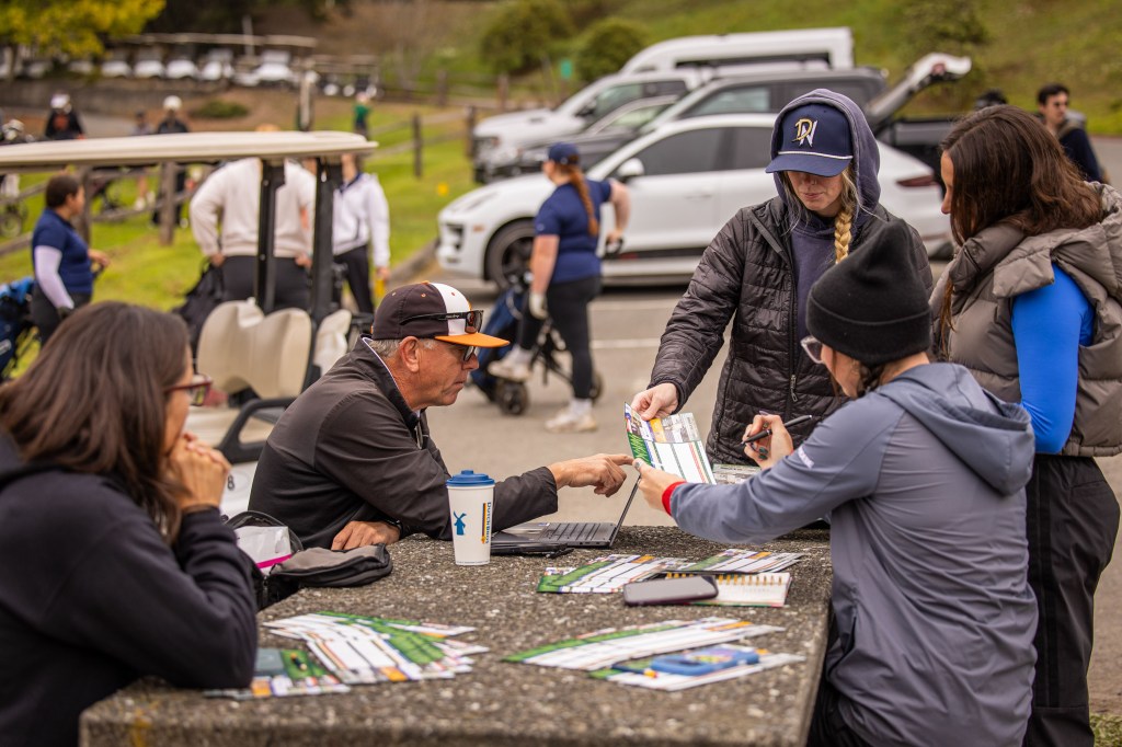 A group of people discussing charts and documents at a table near a golf course, with golf carts and cars in the background.