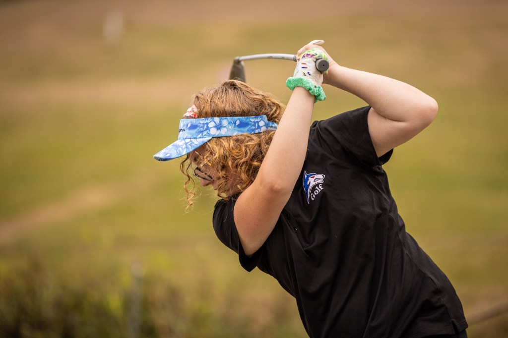A young girl wearing a visor and a black t-shirt swings a golf club on a grassy course.
