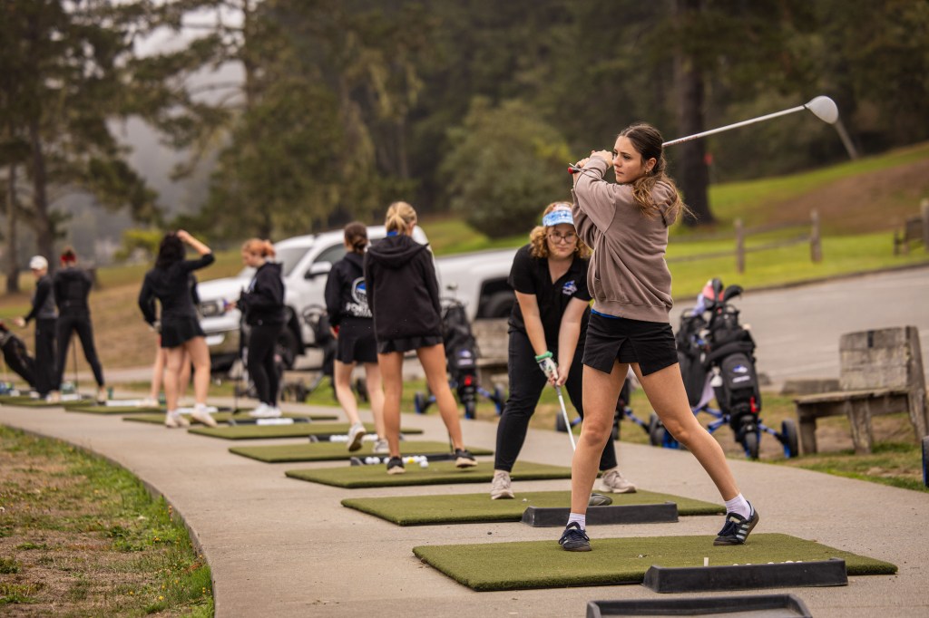 A group of young women practicing golf at a driving range, with one golfer in the foreground preparing to swing her club while others are in the background. The setting is outdoors with trees in the background.
