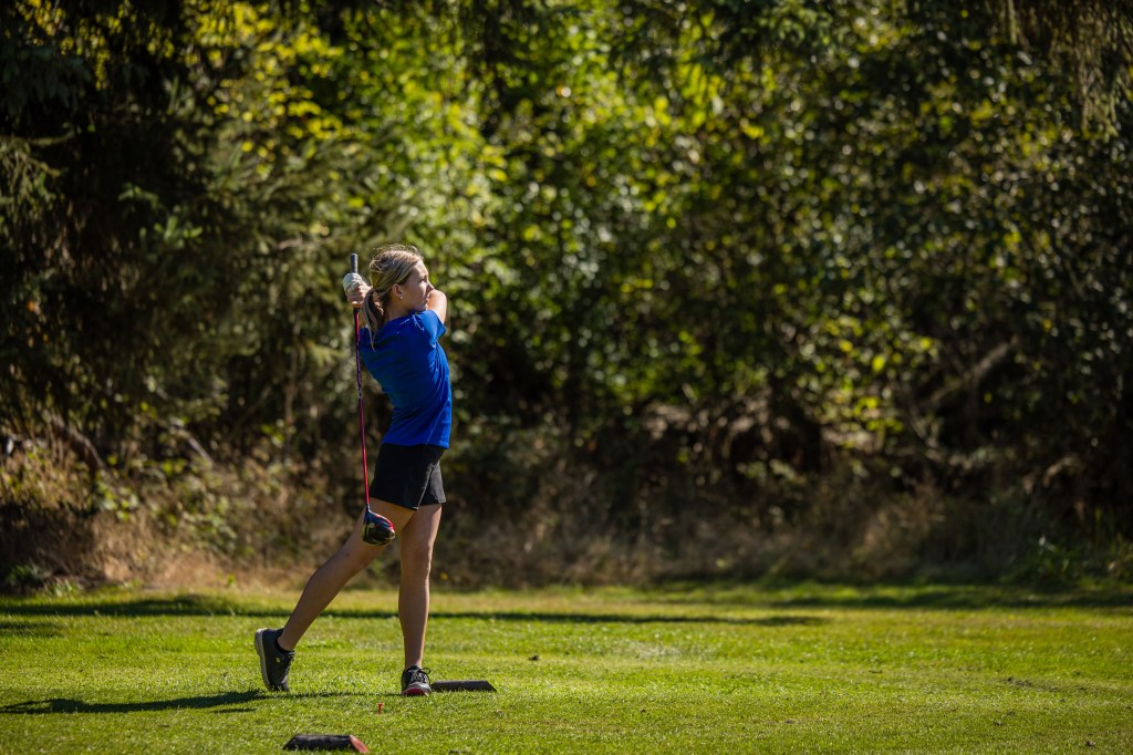 A young girl in a blue shirt swings a golf club on a sunny day, surrounded by greenery.