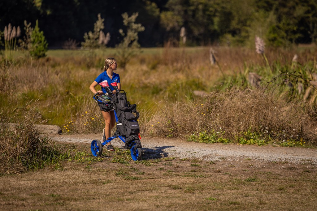 A woman walking with a golf push cart on a scenic path surrounded by tall grass and plants.