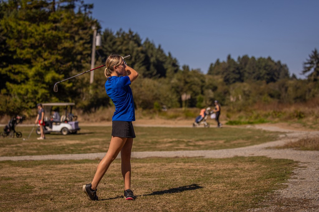 A young female golfer swings a club on a golf course, with trees and other players in the background.