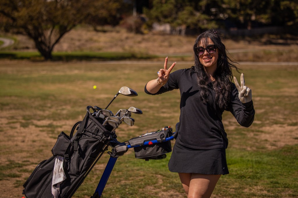 A woman in a black outfit and sunglasses stands on a golf course, smiling and giving a peace sign while holding a golf bag.