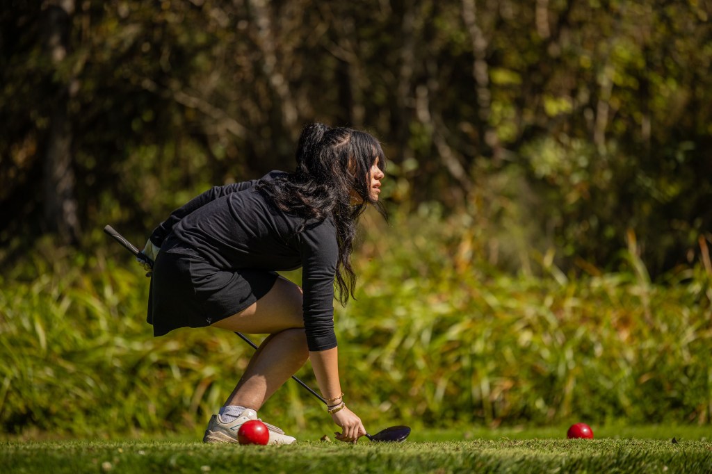 A woman in a black outfit crouches down on a golf course, preparing to hit a golf ball near a red tee.