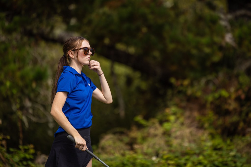 A woman in a blue polo shirt and sunglasses stands on a golf course, thoughtfully looking ahead while holding a golf club.