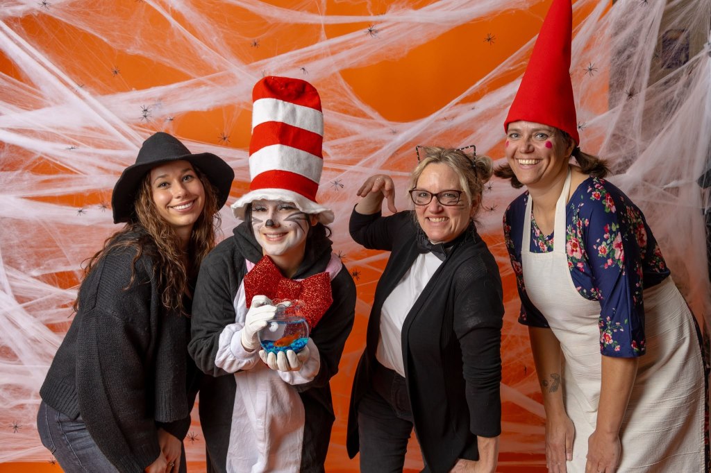 Four women in costume posing together in front of an orange background with spiderweb decorations. One woman is dressed as the Cat in the Hat, another in a witch costume, and two others in playful outfits.