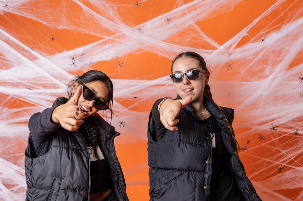 Two women wearing sunglasses and black vests pose playfully with their hands in finger-gun gestures against an orange background decorated with spider webs and small plastic spiders.
