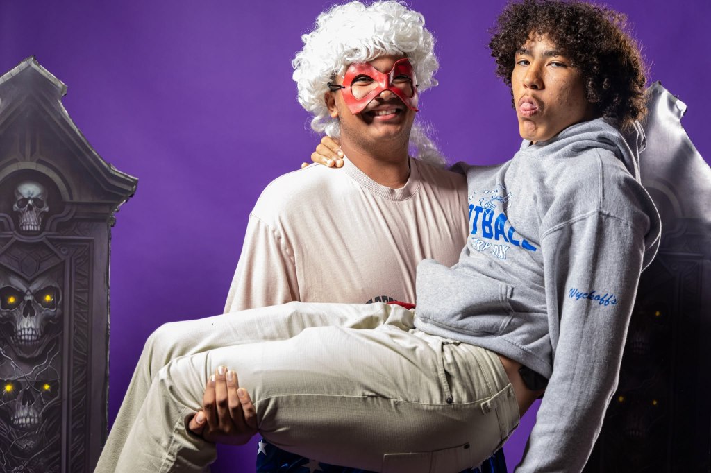 Two people posing for a playful photo, one dressed in a white wig and mask while holding the other in a sweatshirt. The background features gothic-style decorations with skull motifs and a vibrant purple backdrop.
