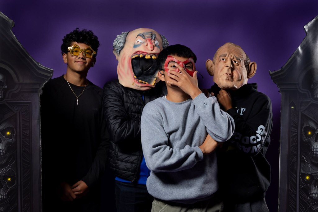 Four individuals wearing various masks and costumes pose in front of a purple backdrop with eerie decorations.