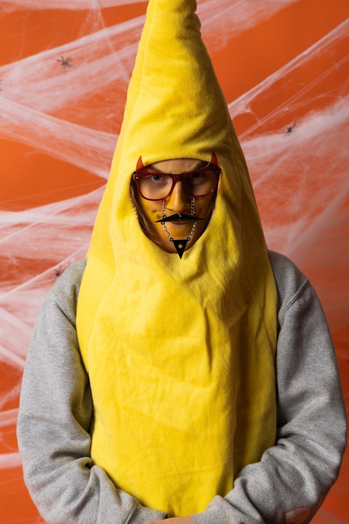 A person dressed in a yellow banana costume with glasses and a unique facial accessory, standing against an orange background decorated with spider web and spiders.
